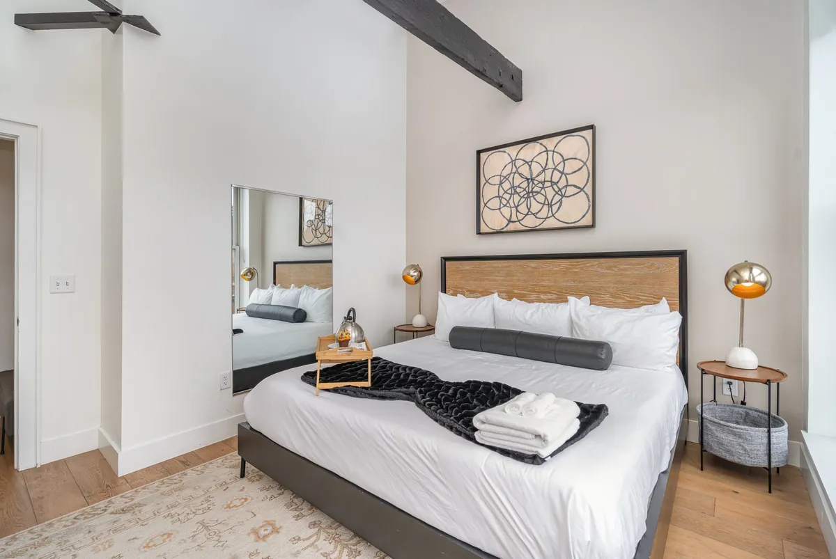 A serene and stylish bedroom with a plush bed, decorative artwork, soft lighting, and a large mirror. The space feels tranquil and comfortable, ideal for restful nights after busy days.