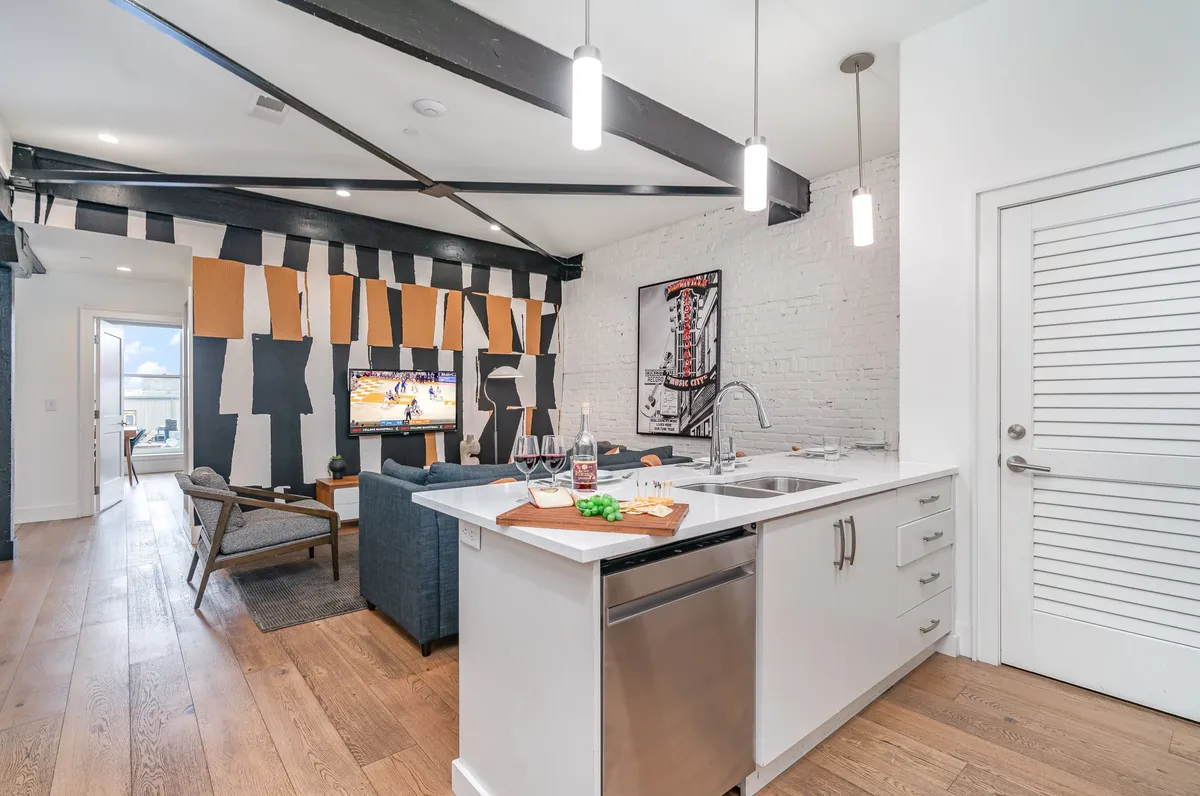 A stylish, open-concept living area featuring a modern kitchen island with a sink, inviting textured wall art, a comfortable seating area, and a television displaying a sports game. The space is lively, with enough room for friends to gather, cook, and relax while watching TV.