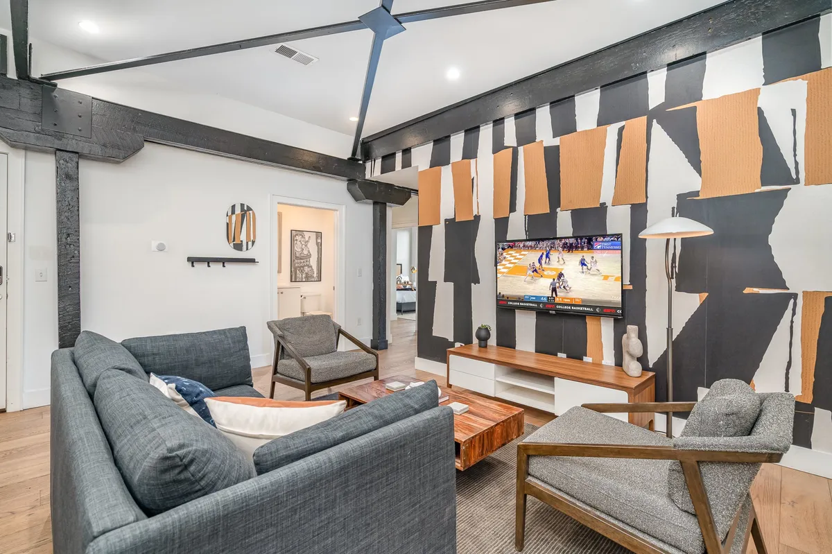 The image showcases a stylish, modern living room with bold abstract wall art, a large flat-screen TV, a sleek wooden coffee table, and inviting modern armchairs arranged for conversation. The atmosphere feels comfortable yet vibrant, inviting guests to relax, socialize, or enjoy a night in.