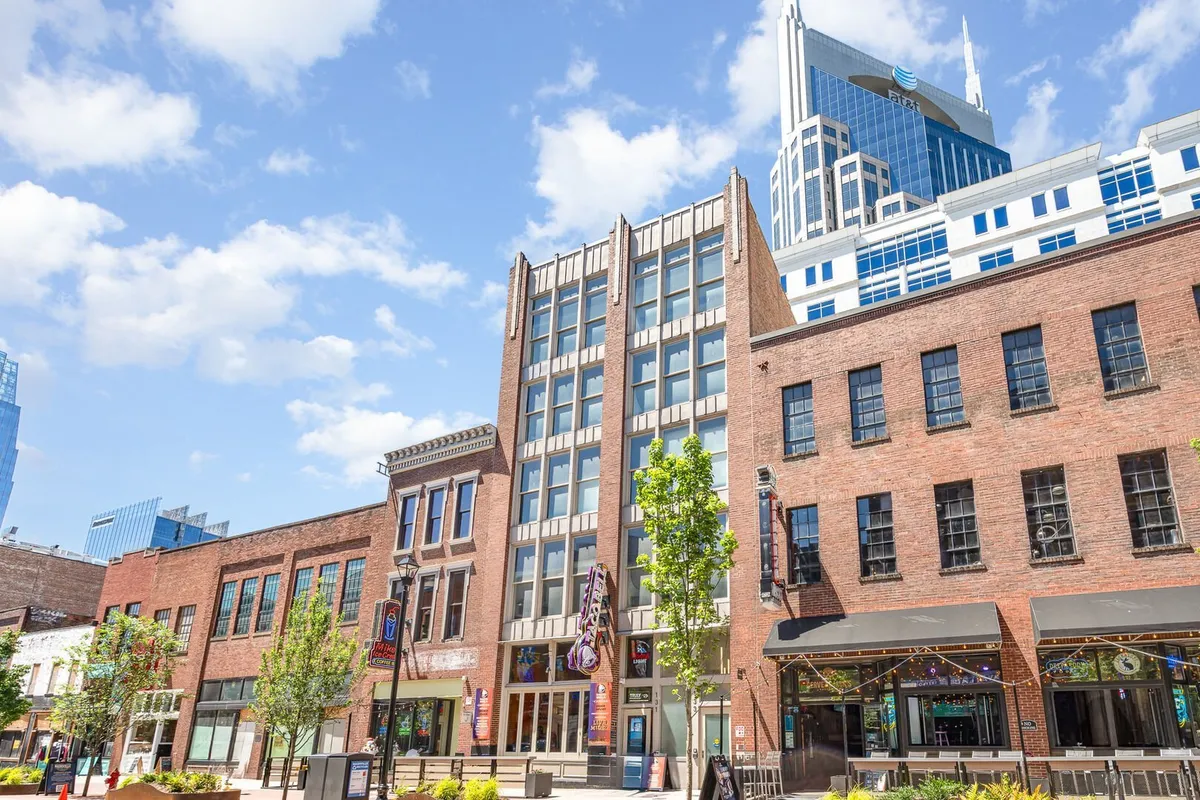 This image captures a lively urban pedestrian area framed by a historic brick building and a modern skyscraper. The scene is filled with people engaged in social activities—shopping, dining, and gathering outdoors—conveying the energy and excitement of downtown life. Outdoor seating and vibrant landscaping reinforce a sense of community celebration.
