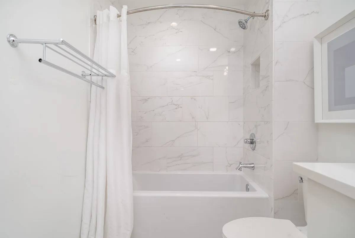 A bright, serene bathroom featuring a combination shower and bathtub with a white curtain and modern marble tiles. A sleek sink and countertop are visible, creating a fresh and inviting space ideal for both quick showers and leisurely soaks.