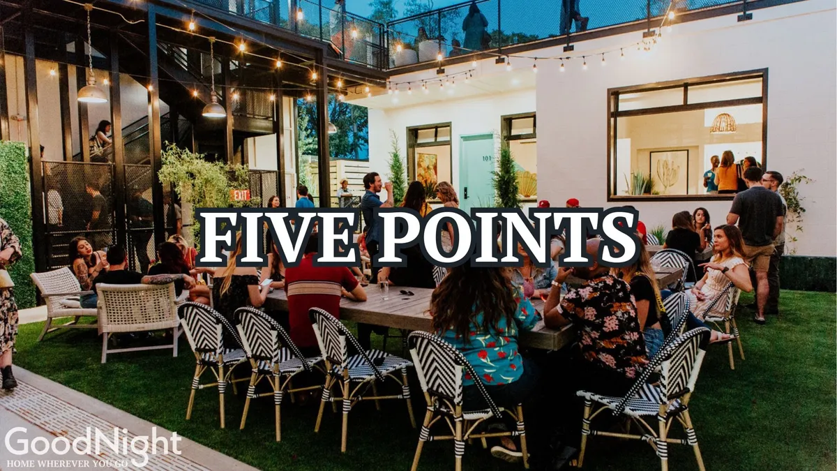 A large outdoor gathering table sits beneath festive string lights. Groups of people are socializing and celebrating against a backdrop of modern architecture. The cheerful ambiance, warm lighting, and communal setting suggest shared meals, toasts, and memorable group moments.
